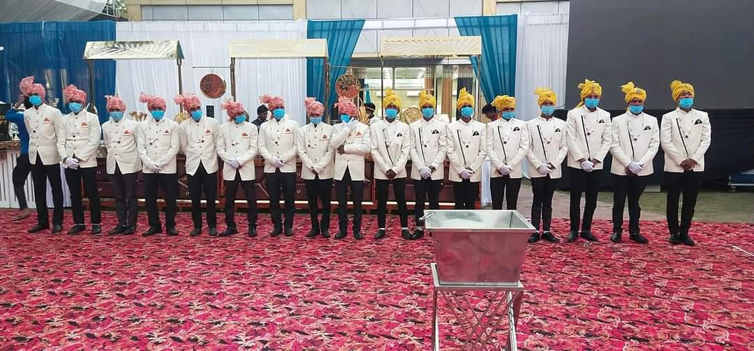 Sixteen waiters in white bandhgala coats and coloured safas standing in a line before service begins