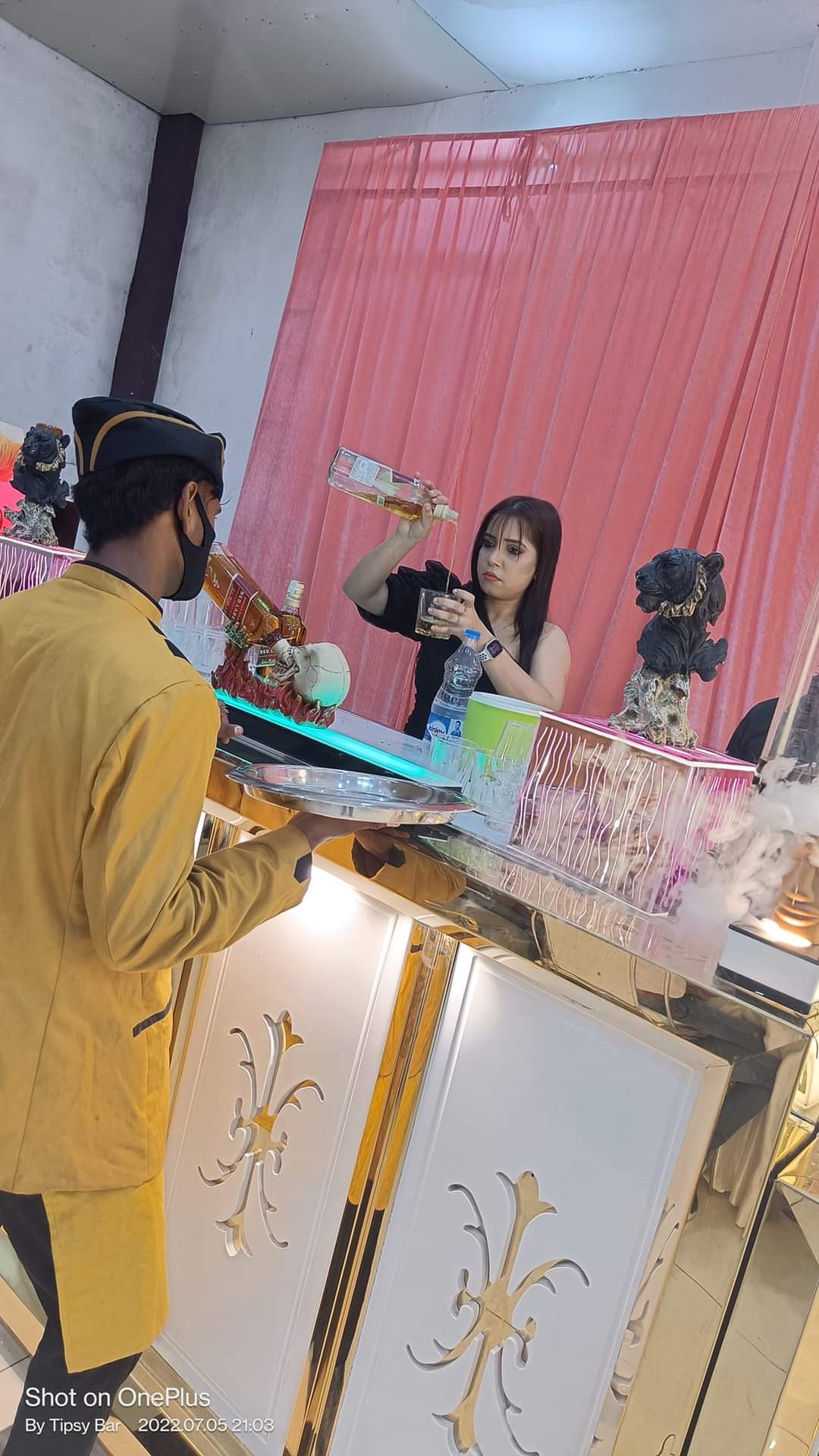 A bartender pouring a drink at the counter while a server waits with a tray